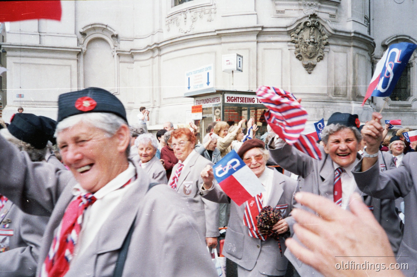 Vintage street scene featuring elderly individuals waving flags and holding signs with "Coca-Cola" logos, likely from the 1960s-1970s. Classic urban architecture with ornate details in background. Crowd appears celebratory, possibly a parade or public event.