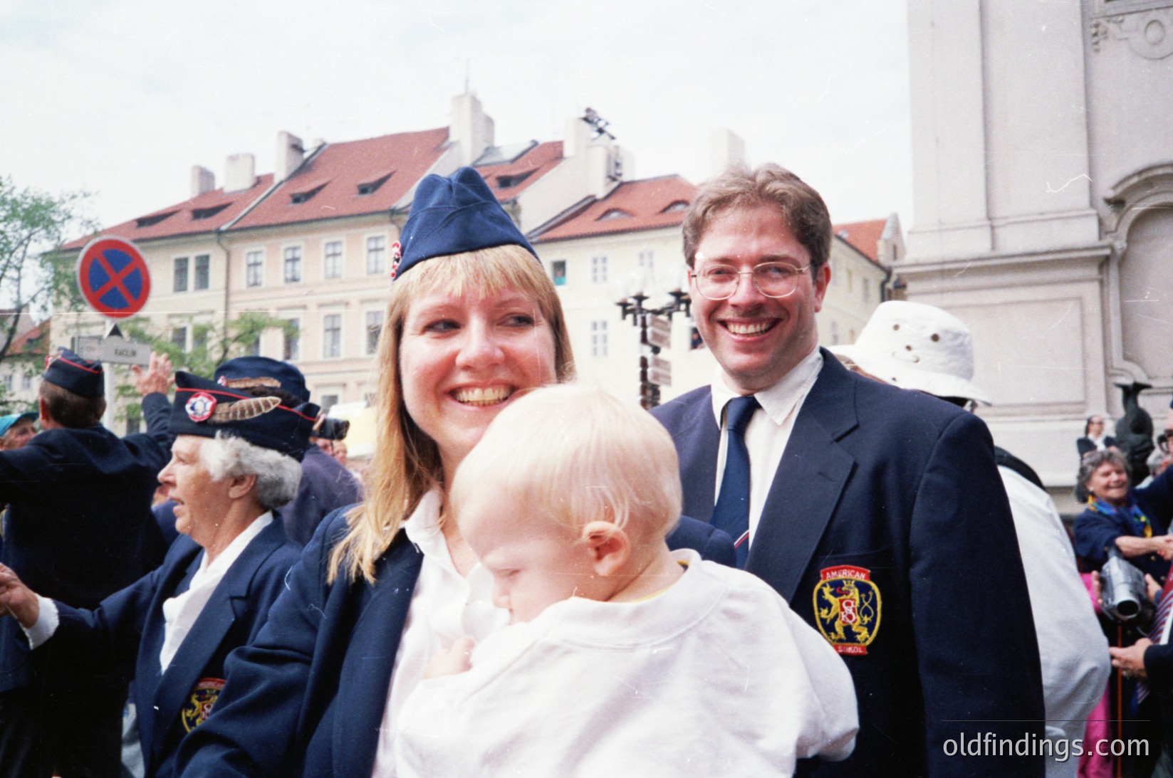 Historic parade scene in Prague’s Old Town, featuring a woman in a blue cap holding a baby, flanked by a man in formal attire with a crest patch. Uniformed participants in traditional Czech folk costumes march in the background. Classic Baroque architecture and cobblestone streets enhance the 1990s-era atmosphere.