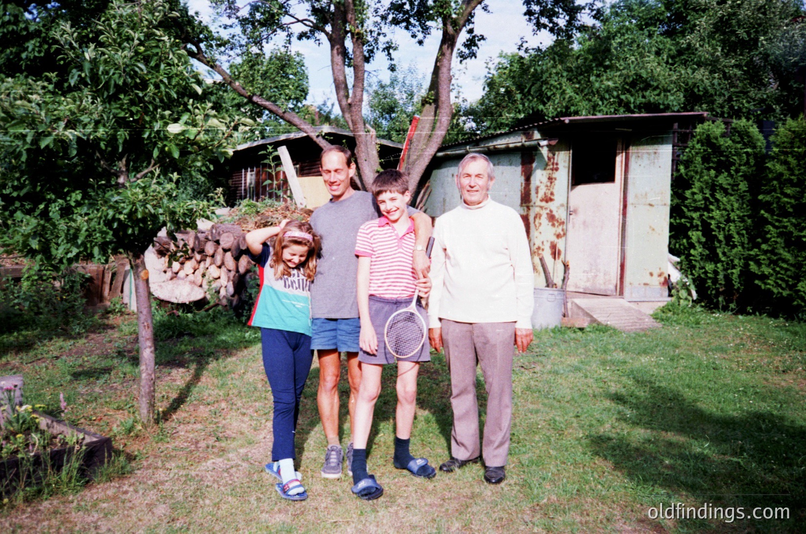 Family portrait in a rural backyard, 1980s–1990s. Three generations pose near a weathered wooden shed and stacked firewood. Adults wear casual, mid-century clothing; the child holds a tennis racket. Lush greenery and overgrown trees suggest a suburban or countryside setting.