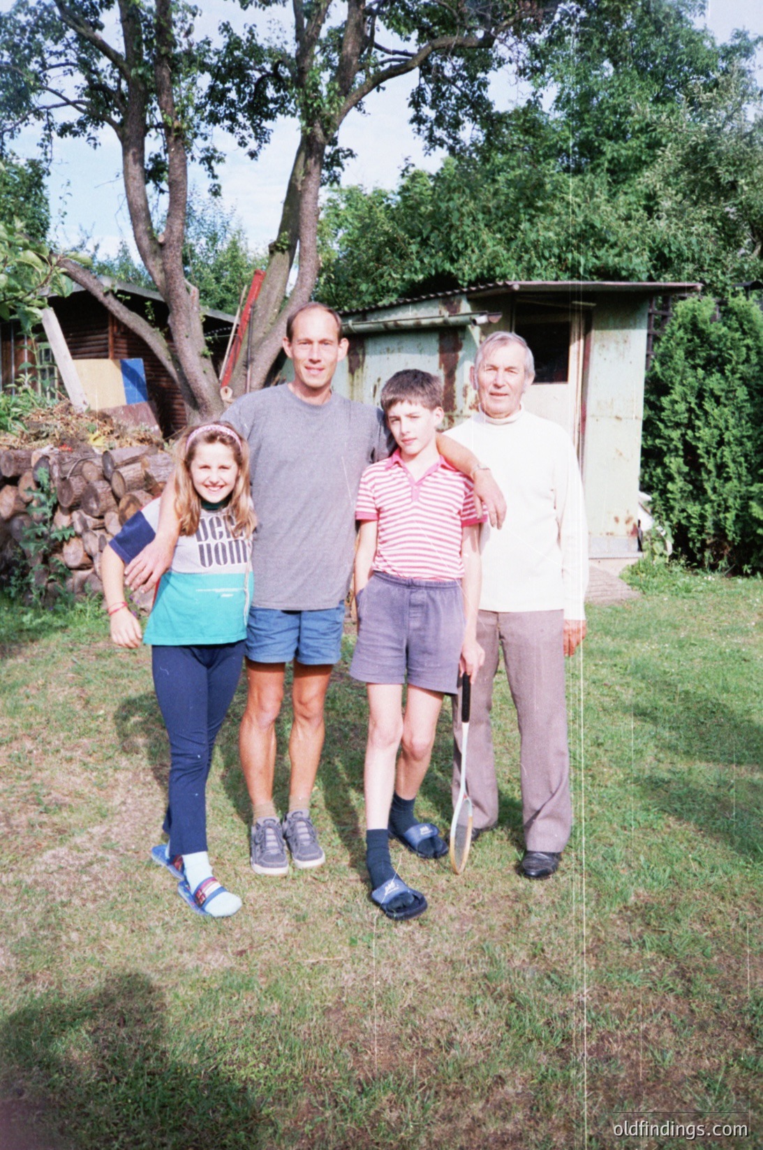 Family portrait in a rural outdoor setting, likely mid-20th century. Four individuals pose in front of a small wooden shed and stone wall. The adult on the left wears a striped sweater and denim shorts; the man in the center wears a short-sleeve button-up shirt and shorts; the boy in the middle wears a striped polo shirt and shorts; the elderly man on the right wears a long-sleeve shirt, shorts, and holds a cane. Lush greenery and trees frame the scene.
