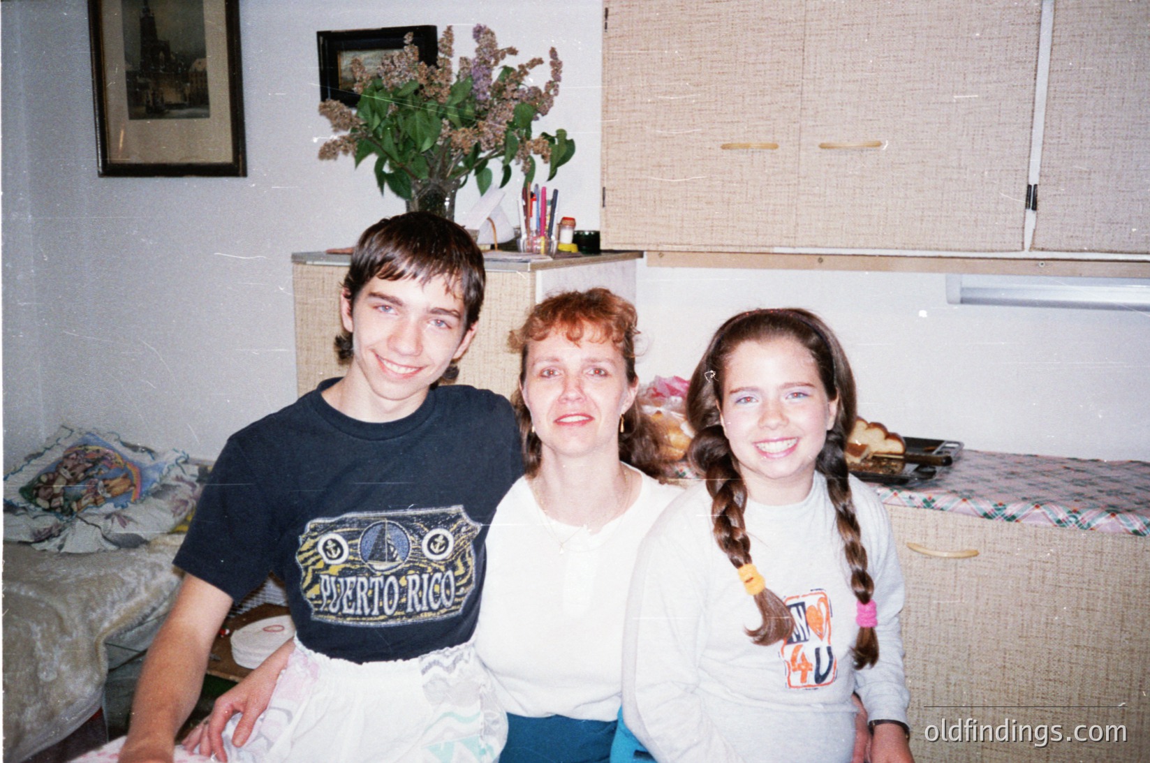 Family portrait from the 1990s featuring a mother and two children in a cozy indoor setting. The boy wears a dark "Puerto Rico" t-shirt, the girl a light "I ♥ U" shirt with braided hair, and the mother a neutral top. Decor includes a floral arrangement, a shelf with art supplies, and a patterned bedspread. Warm, nostalgic home atmosphere.