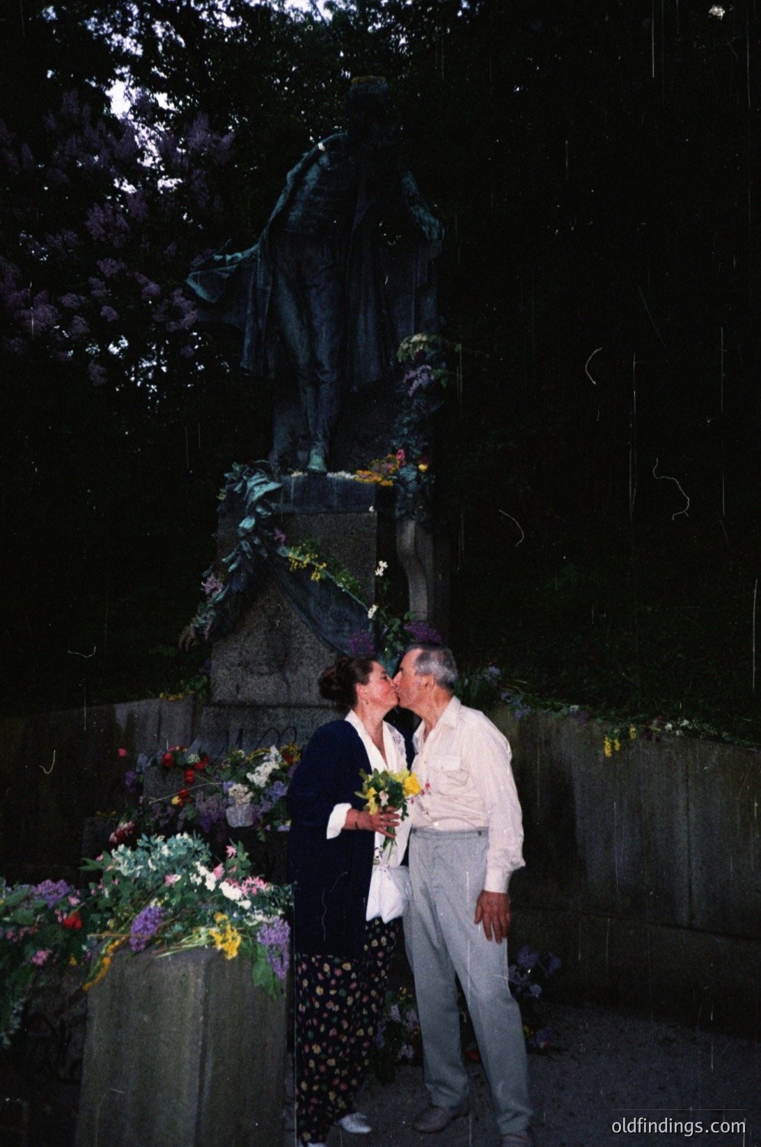 Couple sharing a tender moment at a solemn monument adorned with floral tributes. Dark, dramatic lighting enhances the statue’s silhouette and lush greenery. Mid-20th century attire suggests a formal or commemorative occasion.