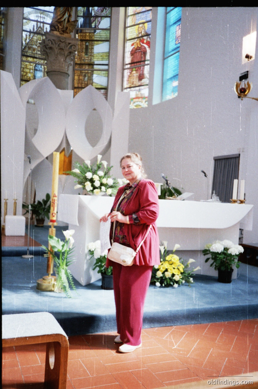 Mid-century modern church interior with abstract white altar sculpture. Woman in burgundy suit and white clutch stands beside a white marble altar adorned with floral arrangements. Stained glass windows and wooden pews enhance the serene atmosphere. Likely 1960s–1970s European church design.