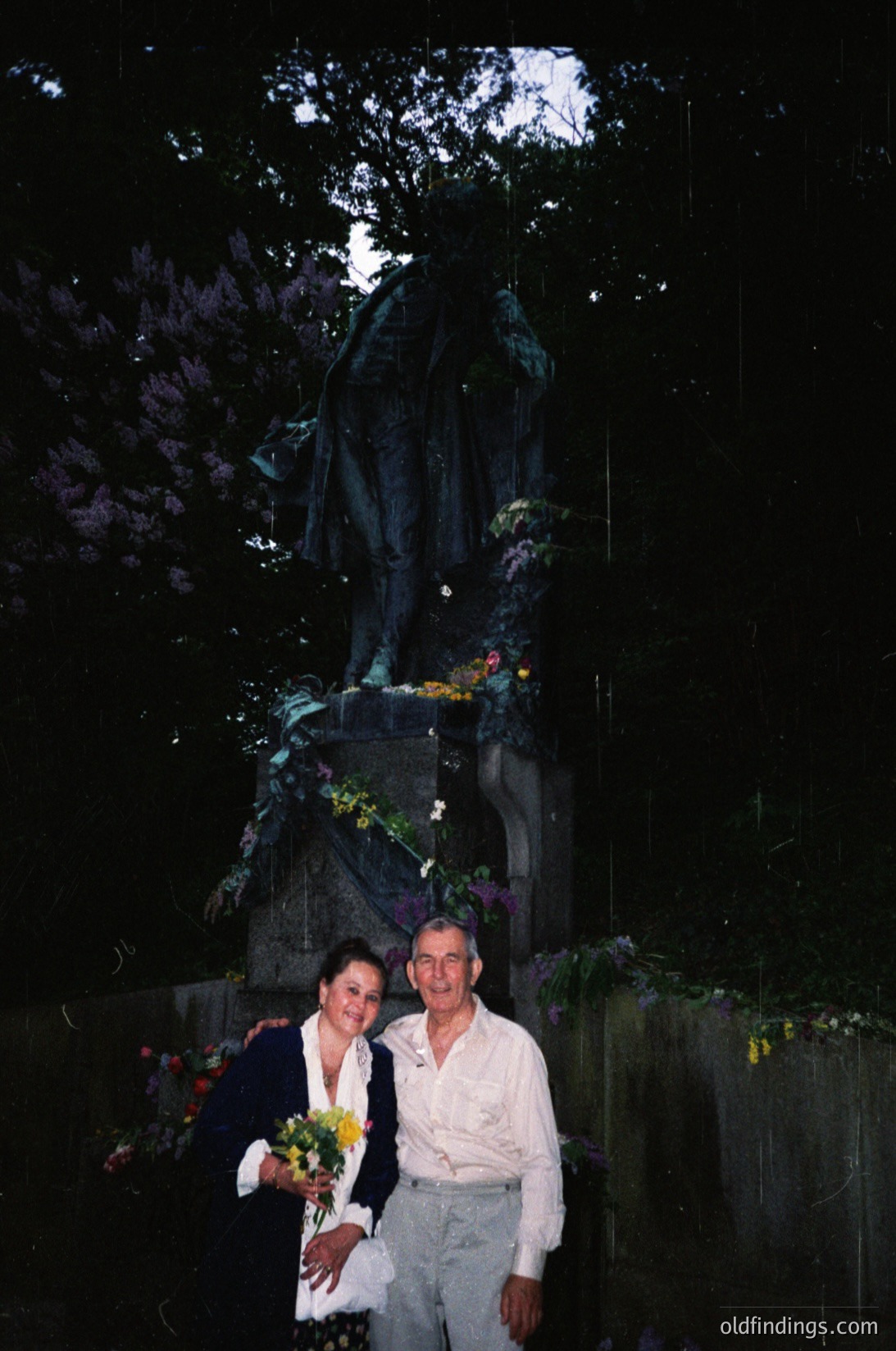 Couple posing beside a dark, moss-covered stone monument with floral tributes, likely a war memorial. Man in light-colored shirt, woman in dark suit holding yellow flowers. Overgrown trees frame the scene, suggesting an outdoor cemetery or memorial park.