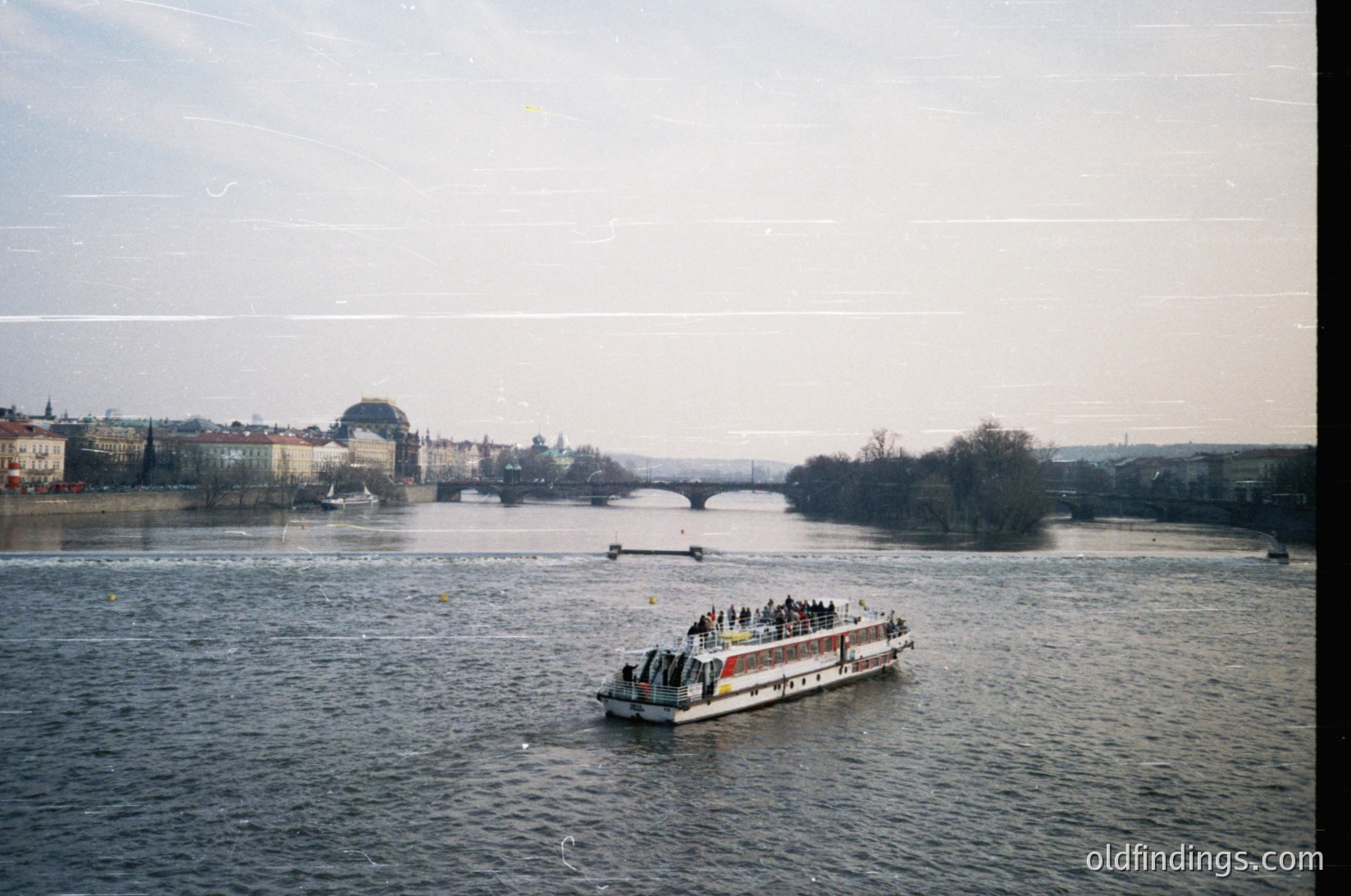 Vintage riverboat with passengers cruising Prague’s Vltava River, framed by historic bridges and Baroque architecture. Overcast skies and muted tones suggest early 20th-century urban tourism.