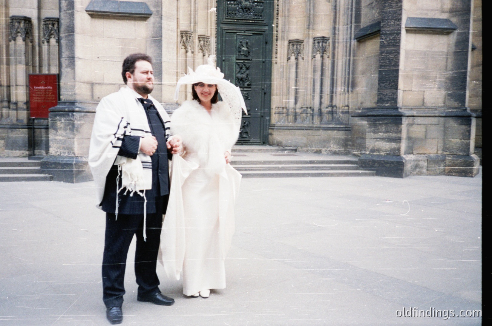 Jewish wedding ceremony in Gothic-style cathedral courtyard. Bride in white veil, groom holding tallit with tefillin. Architectural details include pointed arches and ornate stonework. Likely 1990s–2000s Europe.