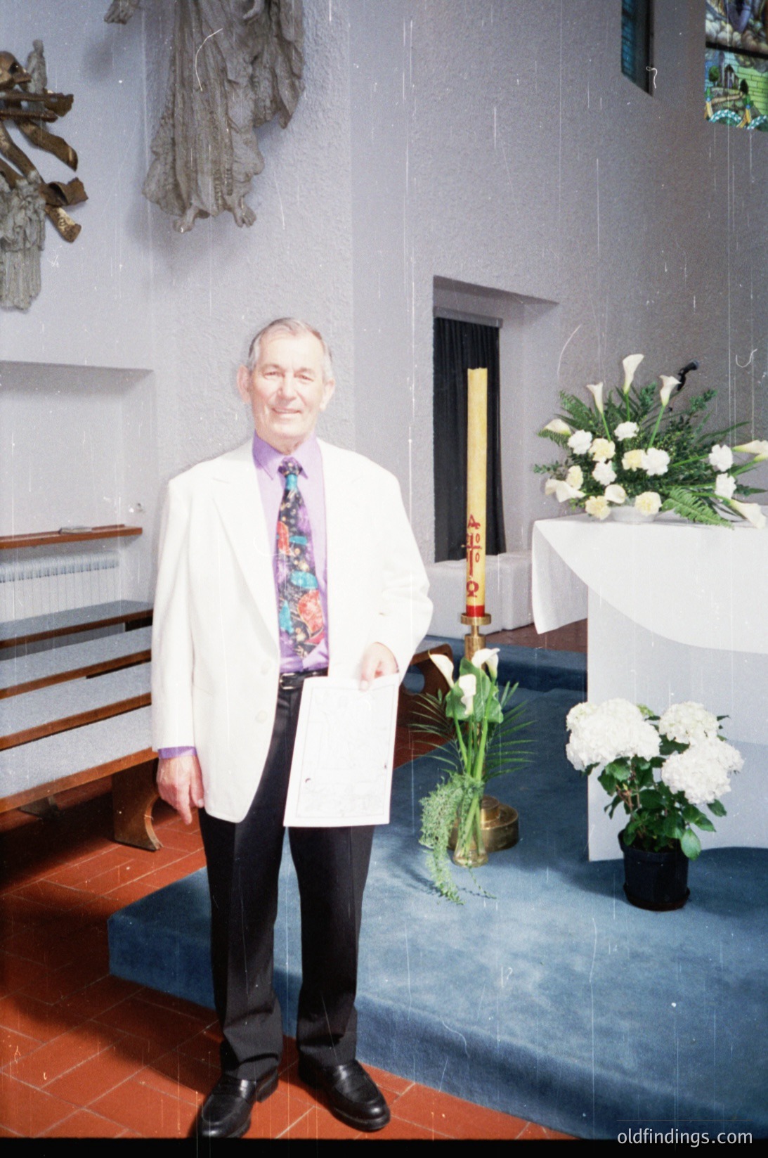Man in white lab coat and patterned tie stands in a minimalist church interior, holding a small object. White altar with floral arrangement and candle holder in background. Red-tiled floor contrasts with light walls. Likely 1990s–2000s, Western Europe/US.