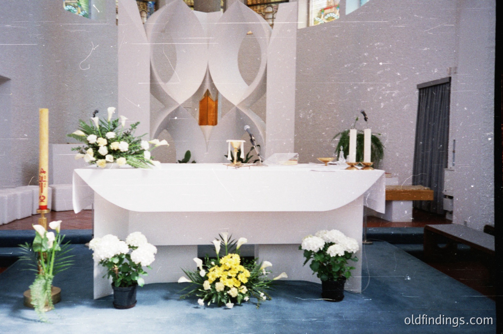 Modernist altar with curved marble altar table, flanked by symmetrical floral arrangements—white lilies and greenery—on black pots. Stained-glass window behind features abstract, wave-like design. Candles and a wooden crucifix on the altar. Mid-century ecclesiastical architecture, likely 1960s–1970s.