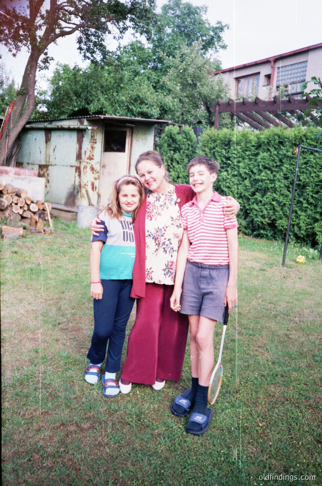 Family portrait in a residential backyard, likely late 20th century. Three individuals pose outdoors near a weathered shed and stacked firewood. The woman wears a floral blouse with a maroon skirt; the girl sports a striped sweater with blue jeans and sneakers. The boy wears a red-and-white striped shirt, shorts, and sneakers. Lush greenery and a wooden fence frame the scene.