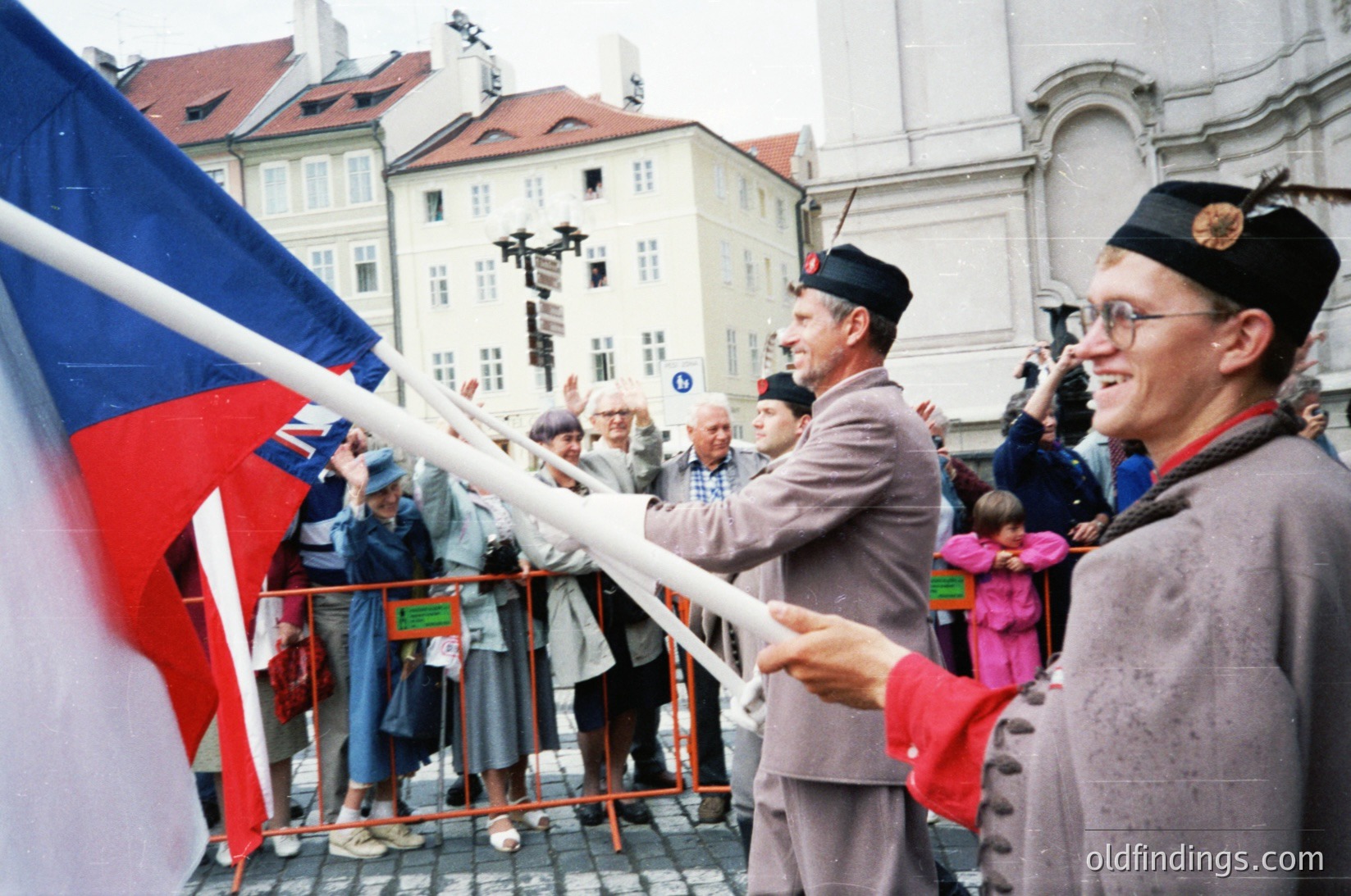 Two men in traditional Czech folk attire—wide-brimmed hats and embroidered vests—hold a large Czech flag during a public gathering. Historic European architecture (baroque-style buildings) and a crowd of onlookers fill the background. Likely a national celebration or patriotic event in Prague,