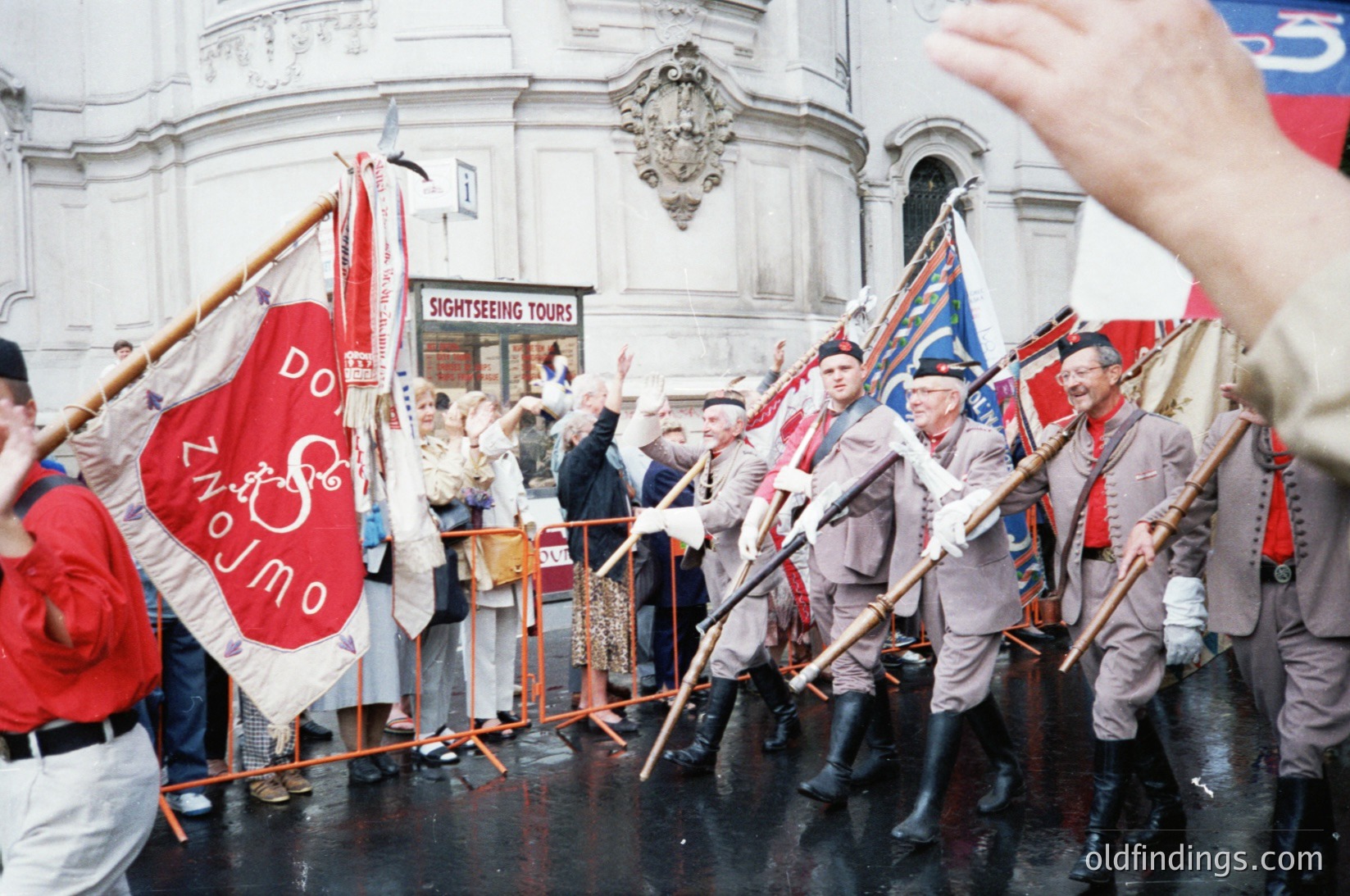 Traditional folk dancers in period costumes perform in a European city square, holding flags with Cyrillic script ("ДОНЖО") and musical instruments. The backdrop features a grand neoclassical building with a "Sightseeing Tours" sign. Likely Bulgaria, 1960s–1980s.