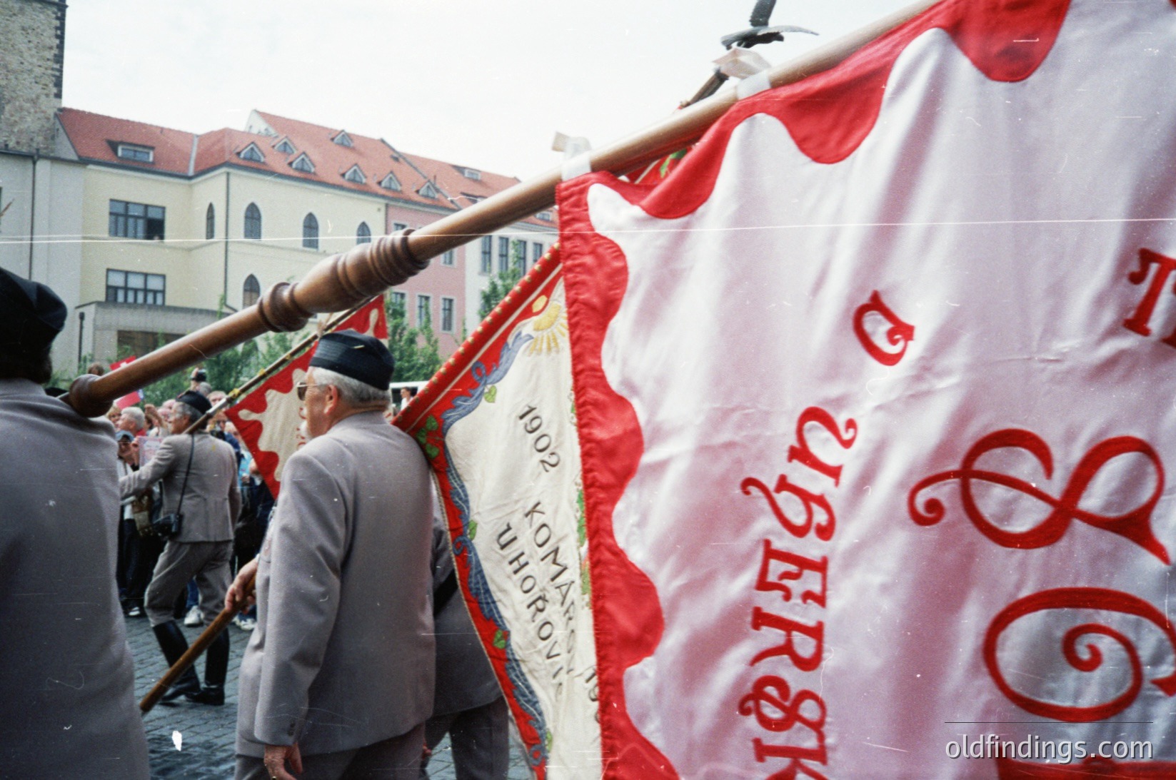 Vintage banner with Cyrillic text ("1902" and "Комсомол") held by elderly men in an urban setting, likely Bulgaria. Red-and-white flag with decorative sun emblem. Crowd in background suggests public event or commemoration.
