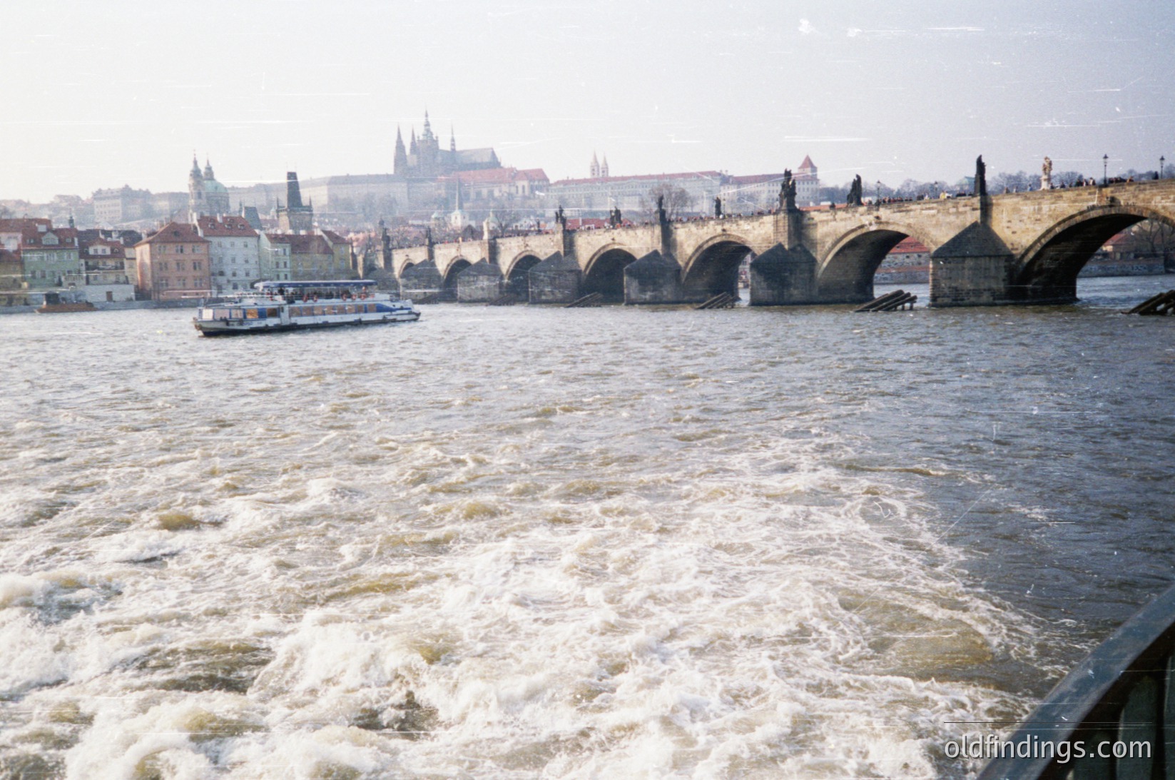 Historic Charles Bridge spanning Prague’s Vltava River, featuring Gothic towers and statues. Classic 1960s/70s color film grain visible. Boat traffic and turbulent water suggest seasonal flow. Iconic Prague skyline in background.