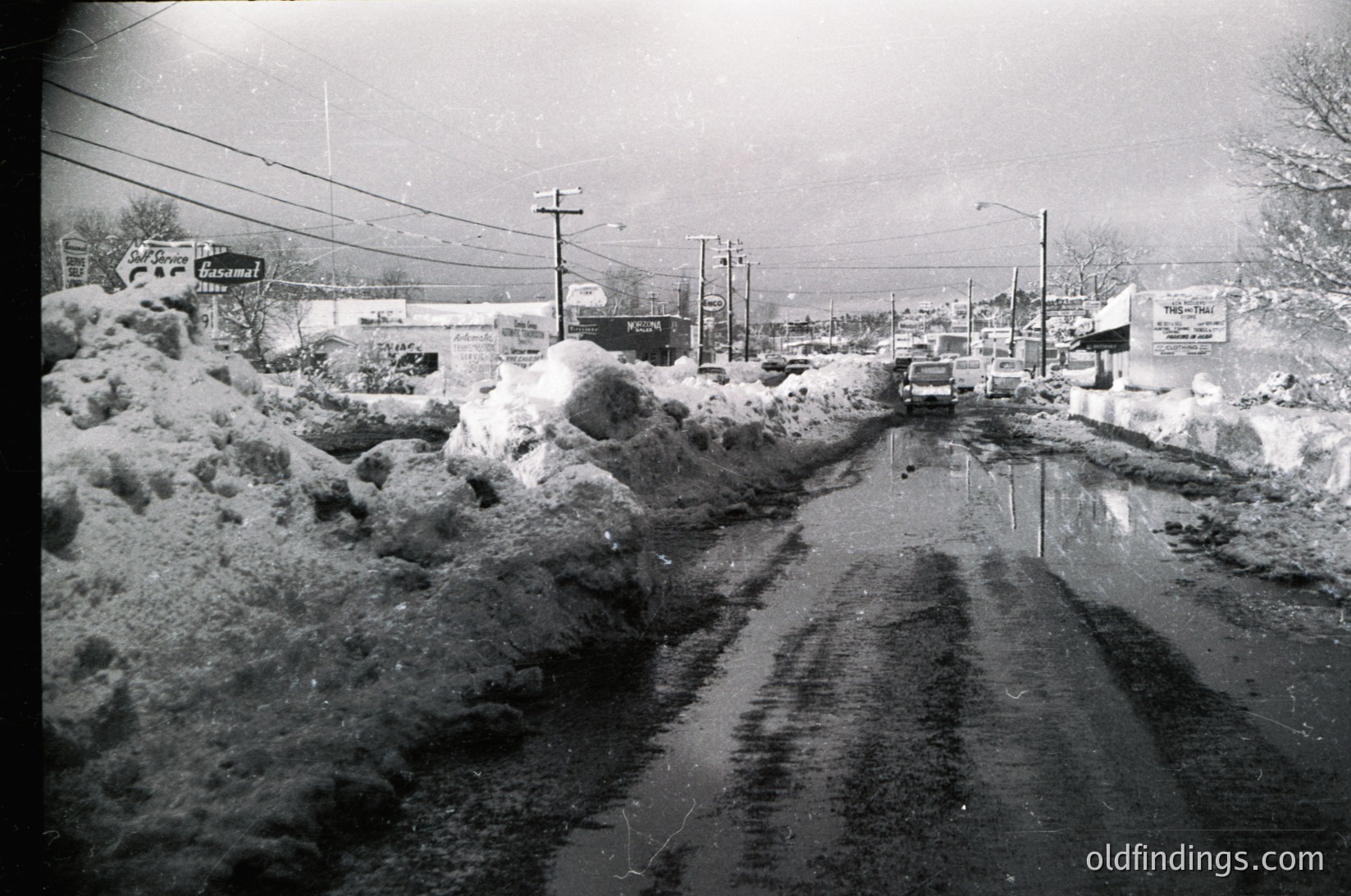 Mid-20th century street blanketed in heavy snow, likely post-1950s. Snowbanks line both sides of a partially cleared road, with a lone car driving through slush. Visible signs include "Vacant" and "Wawa" (convenience store), indicating a small-town American setting. Overhead wires and leafless trees suggest winter conditions.