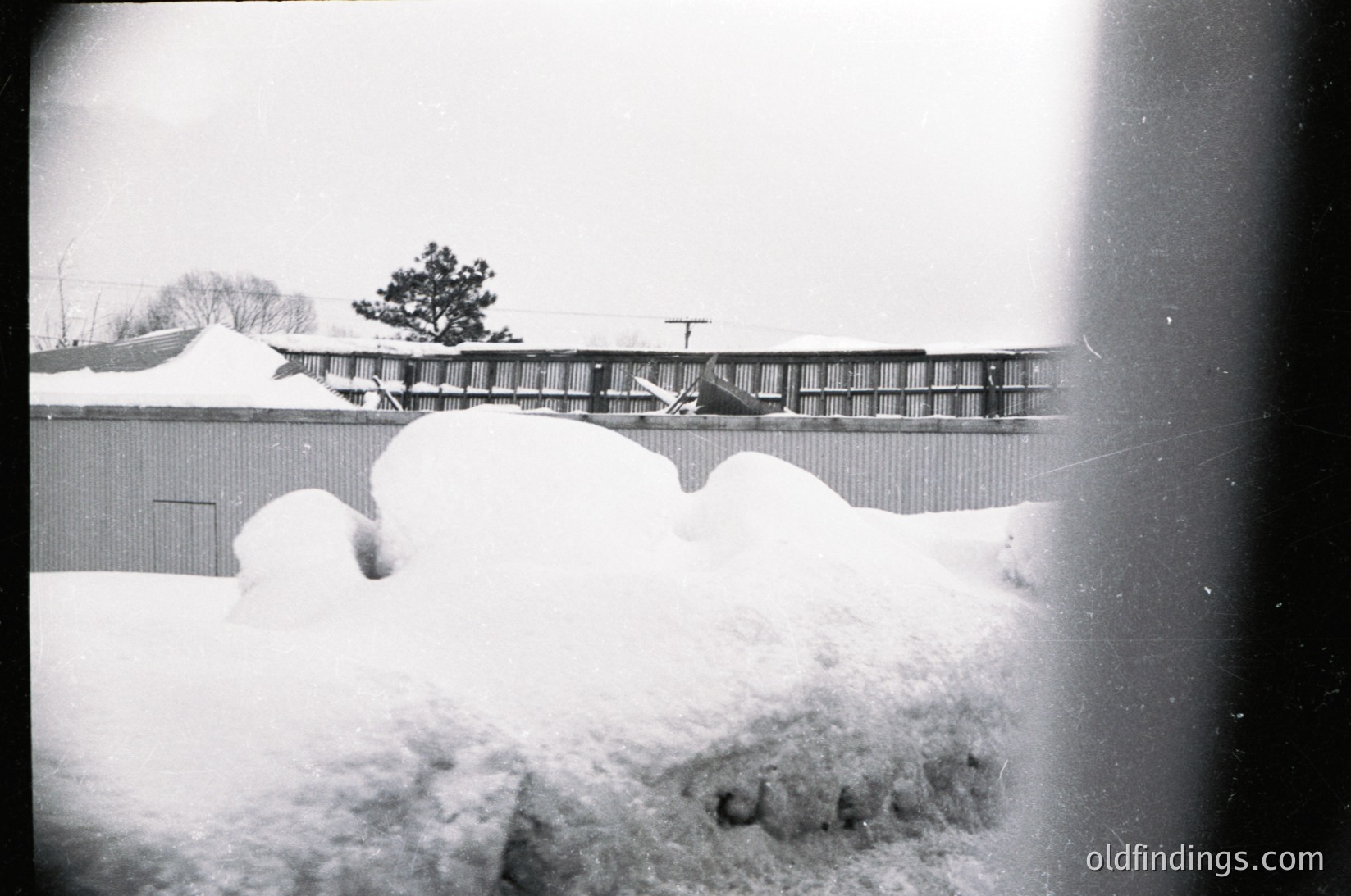 Mid-century industrial scene framed by heavy snowfall, likely 1950s–1960s. Snowdrifts obscure a low-lying building with a corrugated metal roof and a fence topped with barbed wire. Distant train tracks and a lone tree suggest rural or semi-industrial setting.