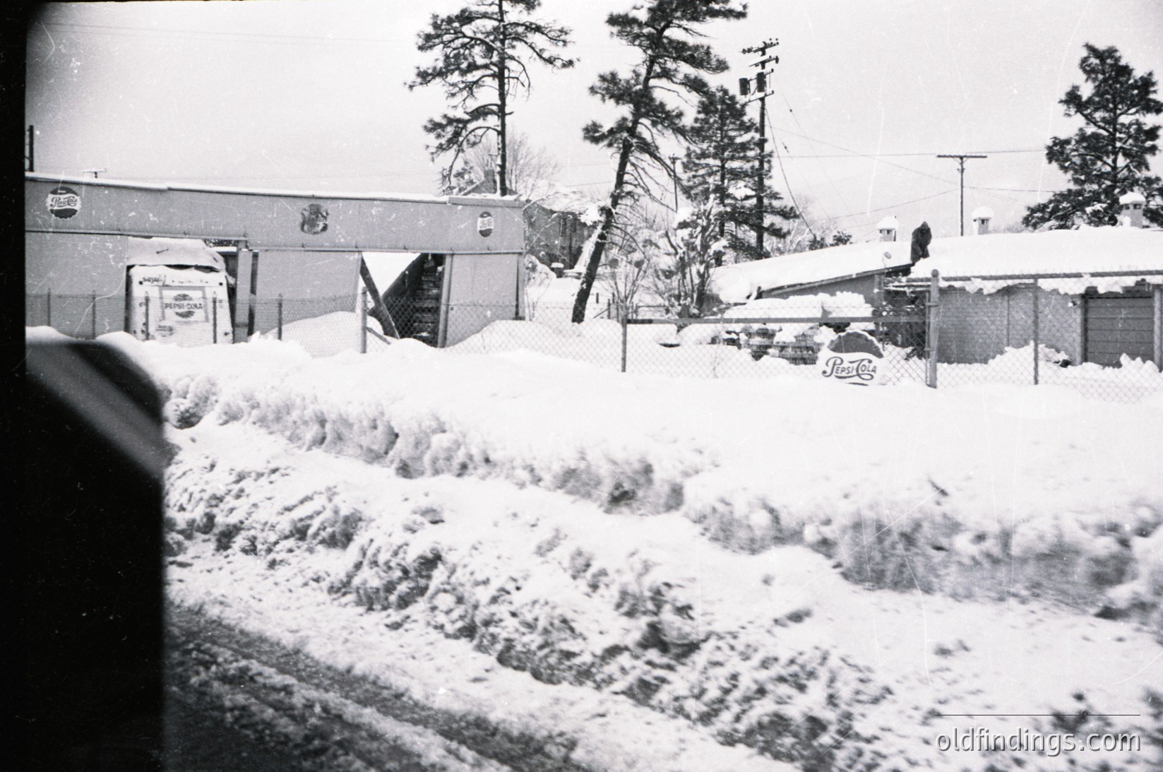 Black-and-white winter scene showing a snow-covered residential area with a partially buried road. Snowdrifts line a street, obscuring a parked car’s rear. A single-story house with a sloped roof and utility poles in the background suggest suburban setting. Snow blankets rooftops, fences, and parked vehicles. Likely mid-20th century due to architectural style and monochrome.
