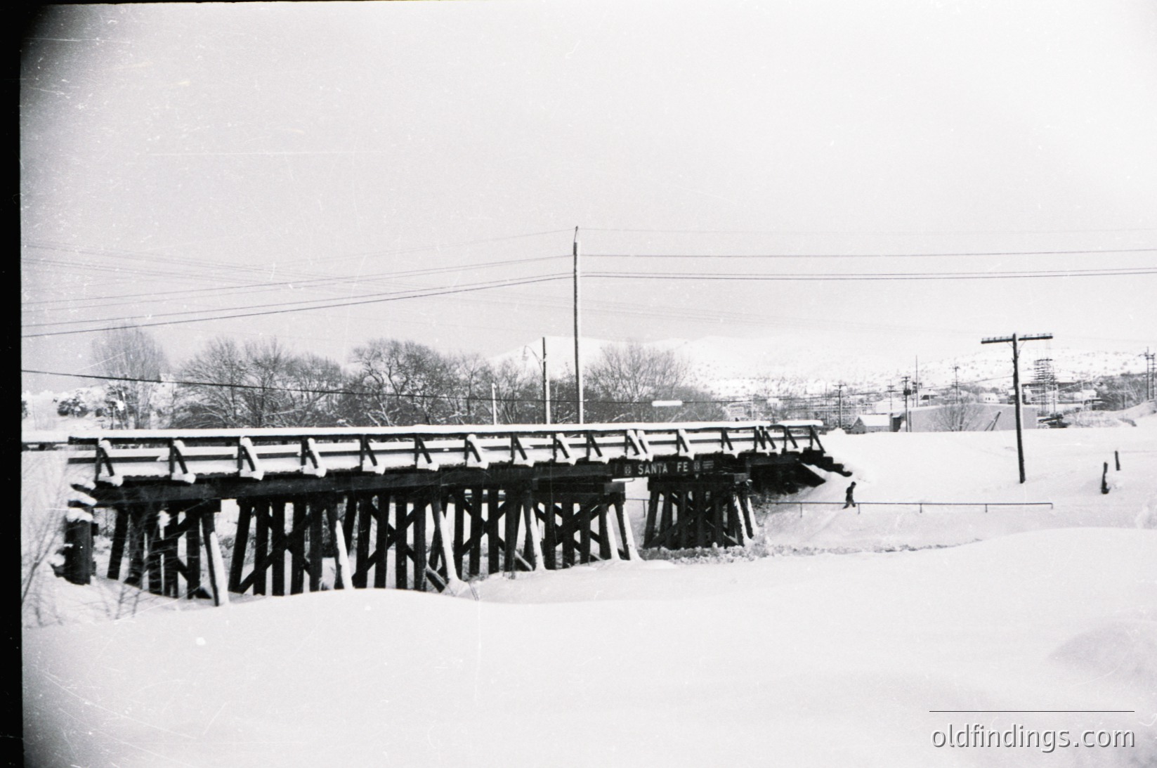 Black-and-white winter scene of a wooden truss bridge spanning a snow-covered stream. Snow blankets ground, trees, and bridge supports, with utility poles and wires extending into the distance. Mid-20th century rural infrastructure, likely North America.