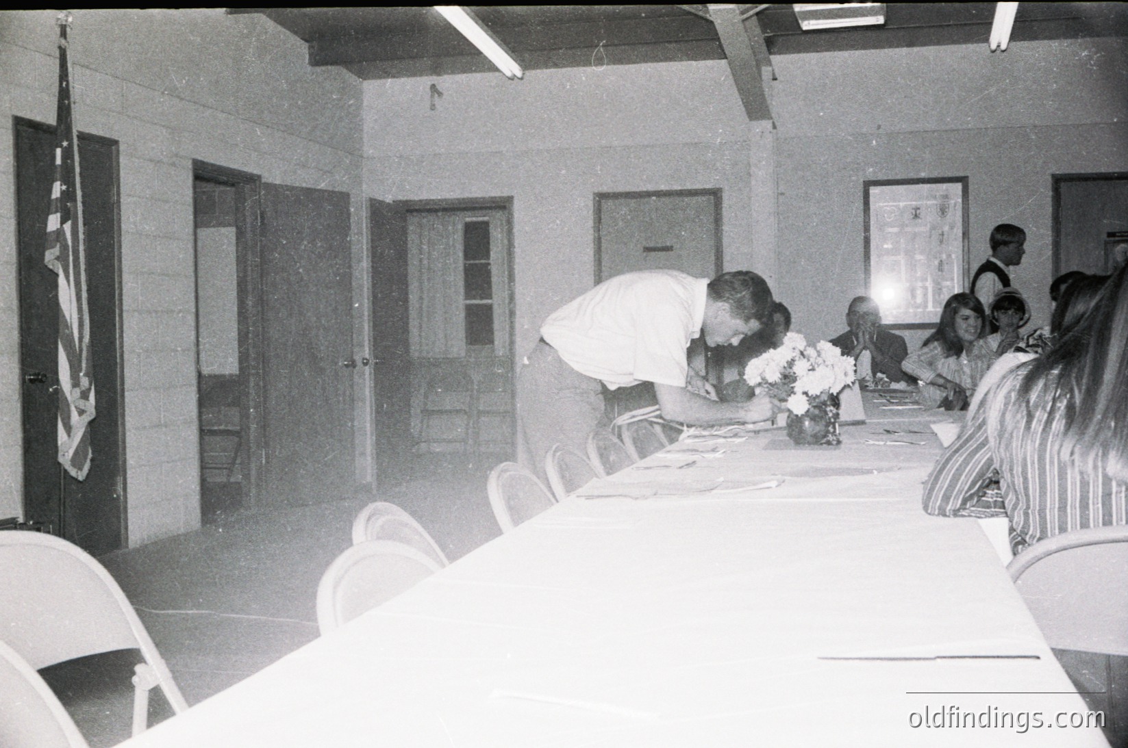 Mid-century indoor gathering with floral centerpiece on a long table. A man in a white shirt bends to place a wreath on a plaque or podium. Attendees in casual 1960s-70s attire sit on plastic chairs. American flag on left wall, framed art on right. Institutional or community hall setting.