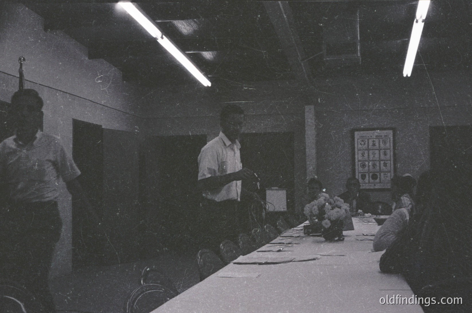 Indoor classroom scene featuring a teacher addressing seated students at a long table. Fluorescent lighting and a chalkboard with Cyrillic letters suggest Eastern European setting. Mid-20th century educational environment, likely –.