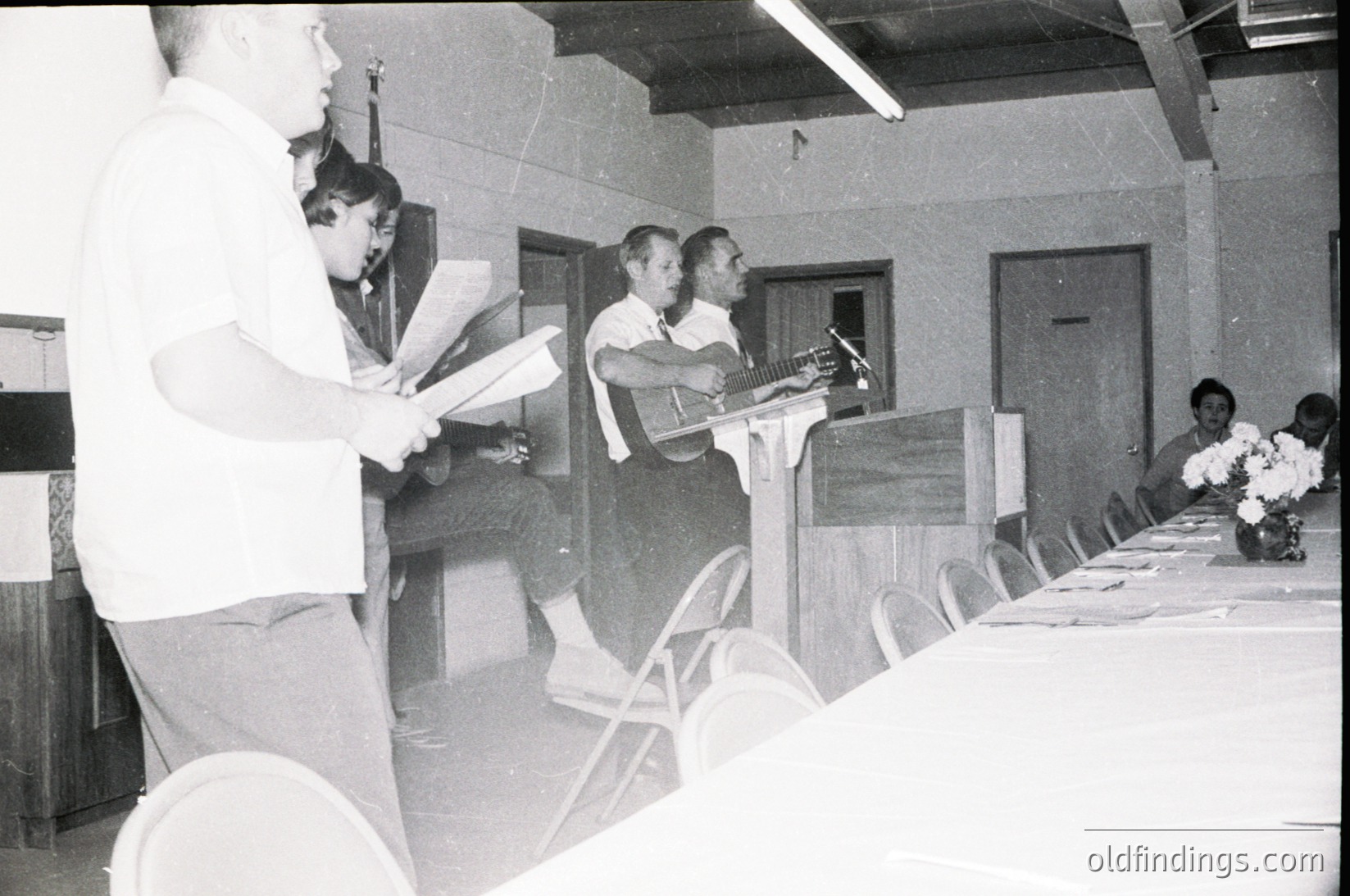 Black-and-white indoor shot of a formal event in the 1960s–70s, featuring a podium with a speaker addressing an audience. Mid-century conference hall with tiered seating, floral centerpieces, and a microphone stand. Attendees in formal attire, including suits and dresses.