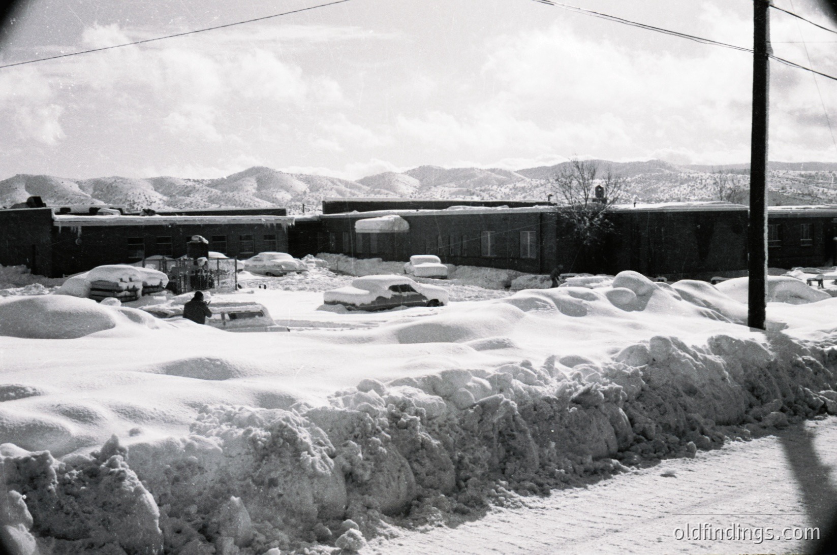Snow-covered urban street with buried vehicles, mid-20th century architecture. Heavy snowbanks line the roadside, partially obscuring a low-rise building with flat roof and utility lines. Snow-laden trees and distant mountains add depth. Likely – winter scene.