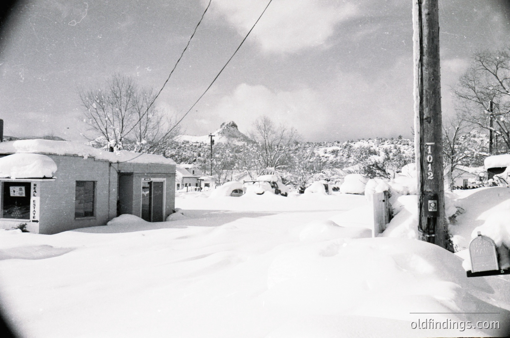 Snow-covered residential street with mid-century concrete block houses, blanketed in winter. Bare trees and utility poles line the road, leading to a distant mountain range under overcast skies. Likely 1950s–1970s U.S. suburban scene.