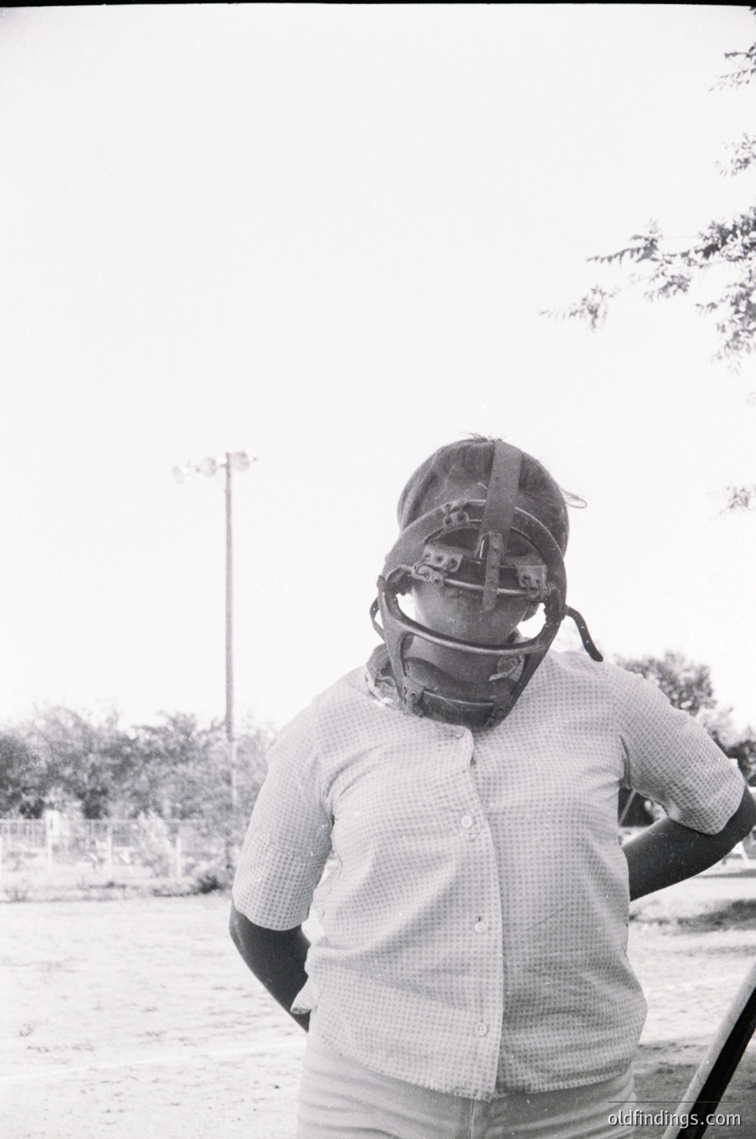 Baseball catcher in vintage mask and checkered shirt, standing outdoors. Classic 1960s-1970s sportswear and protective gear. Likely midwestern U.S. due to landscape and attire.
