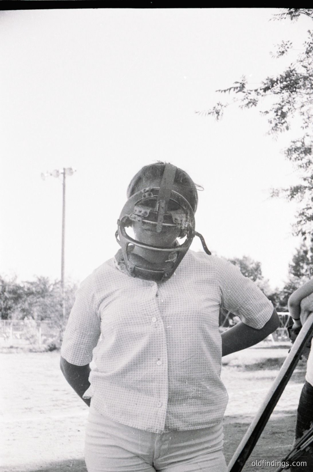 Baseball catcher in vintage protective gear, likely mid-20th century. Checkered shirt and knee-high socks suggest 1950s–1960s style. Outdoor setting with blurred greenery and asphalt, indicating a field or park. Ideal for sports history, vintage sportswear research, or nostalgic stock imagery.