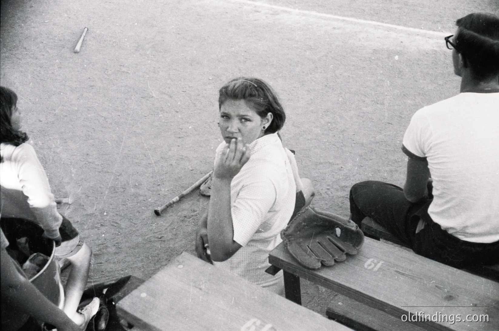 Baseball dugout scene featuring a woman in mid-1960s attire—short-sleeve blouse, skirt, and finger on lip—sitting beside a catcher’s mitt. Faded black-and-white photo captures candid expressions and vintage stadium setting.