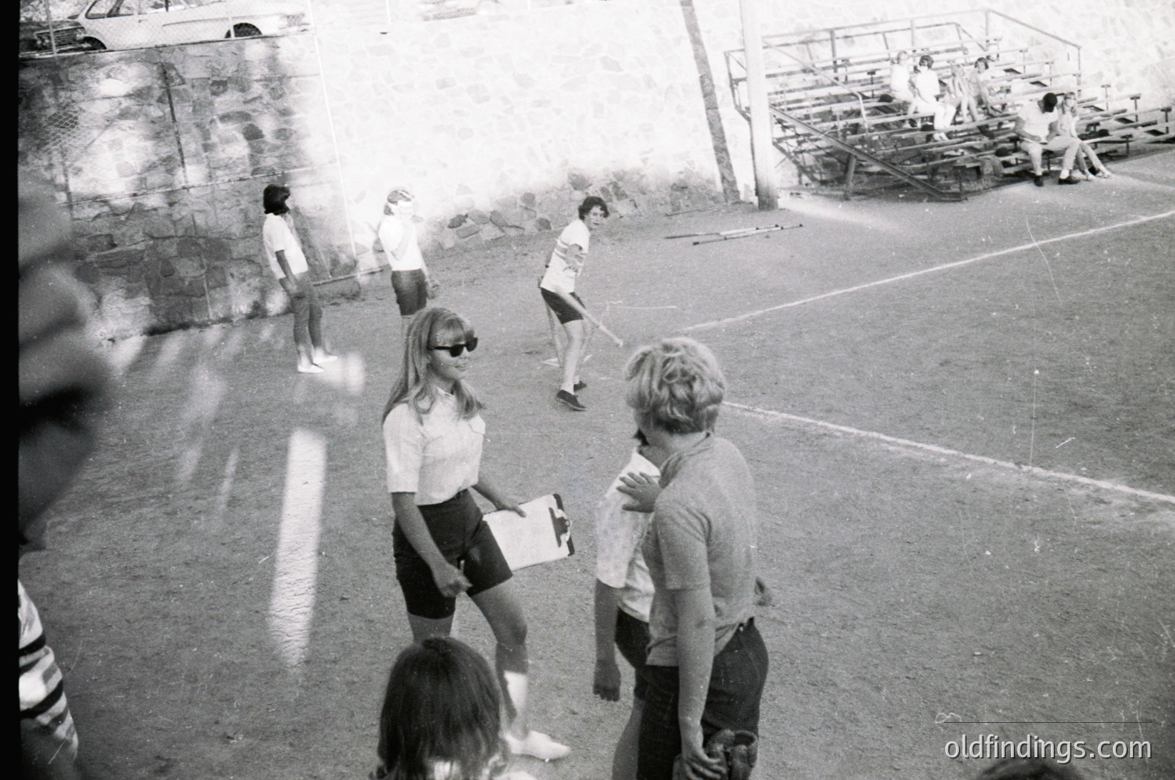 1970s outdoor basketball game in a concrete court setting. Players in short-sleeve shirts and shorts, with one holding a clipboard. Empty bleachers and a fence in background. Casual, informal atmosphere.
