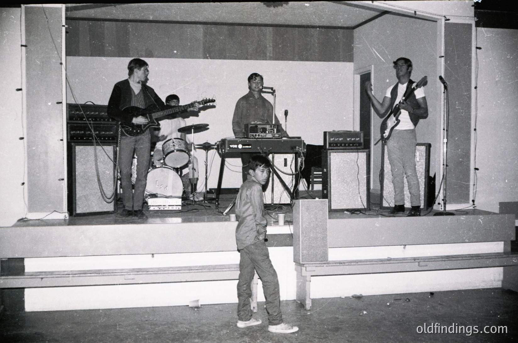 Vintage live band performance on a modest stage with four musicians: electric guitar, keyboard, drums, and bass. Child in foreground, likely a fan, stands on concrete steps. Stage lighting and equipment suggest 1970s–1980s era. Casual attire and minimalist setup reflect underground or community venue vibe.