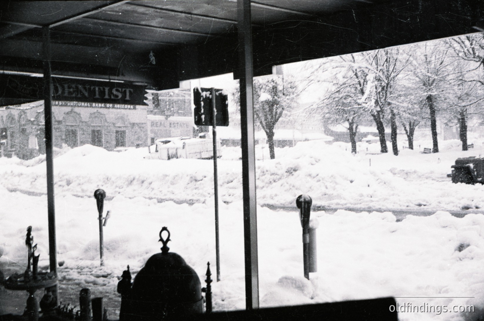 Snow-covered urban street corner with vintage signage: "DENTIST" (International Bank of Austria) and "REAL ESTATE" visible. Black-and-white photo captures mid-20th century (1950s-60s) European cityscape. Snow blankets sidewalks, parked cars, and traffic lights. Distinctive street lamps and barren trees frame the scene.