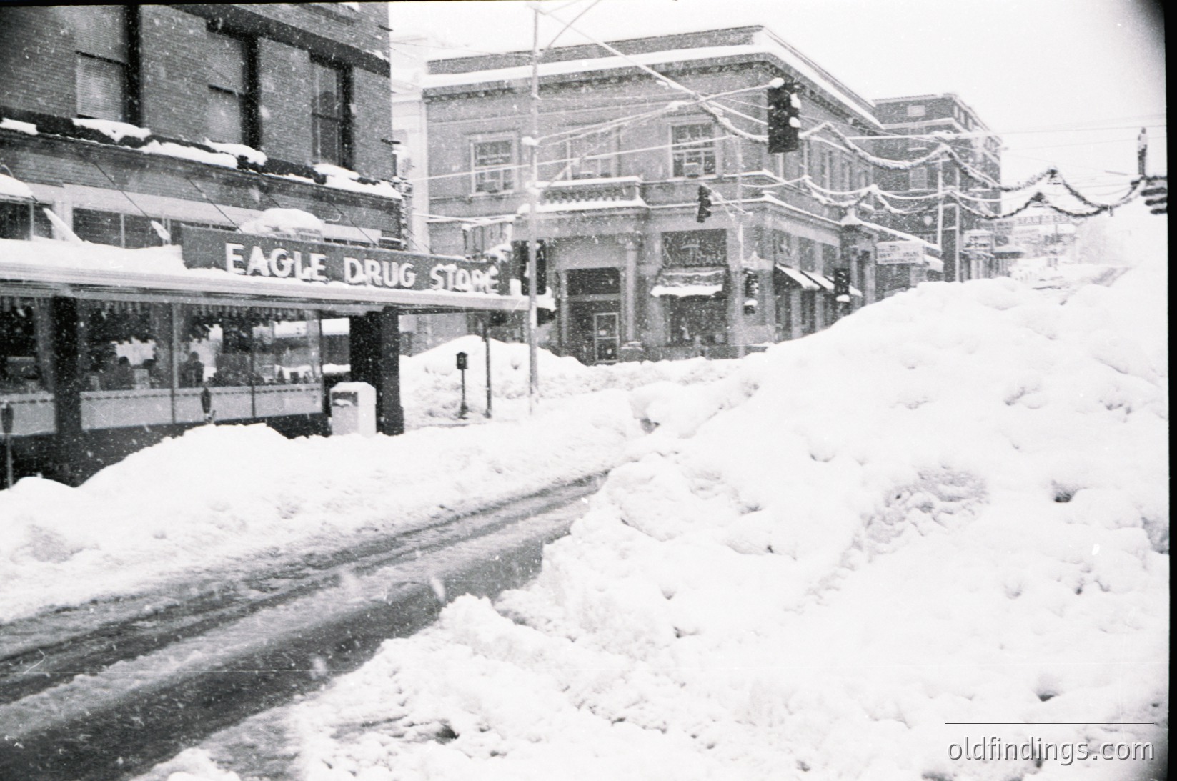 Mid-20th century street scene blanketed in snow, featuring the **Eagle Drug Store** with Art Deco signage. Snow-covered buildings, traffic lights, and sidewalks suggest a winter storm in an urban setting. Likely **1940s–1960s**, North American city.