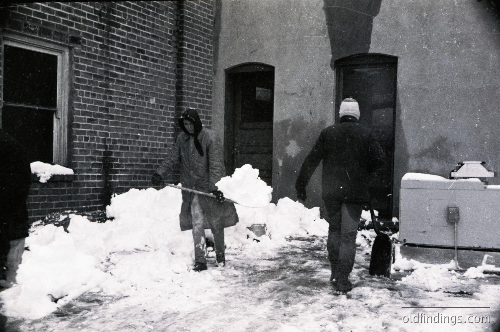 Two men clear snow from a brick-paved alleyway in mid-20th century, likely 1950s–1960s. One shovels snow into a pile, the other pushes a wheelbarrow. Brick buildings and a vintage icebox (refrigerator) visible. Urban winter scene.