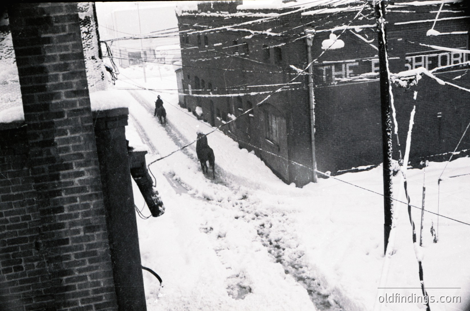 Mid-20th century urban alleyway blanketed in snow, with three workers clearing paths using shovels. Brick buildings flank the scene, including a visible "The Safeway" sign. Overhead wires and bundled snowbanks highlight winter conditions. Likely North American setting, 1950s–1960s.