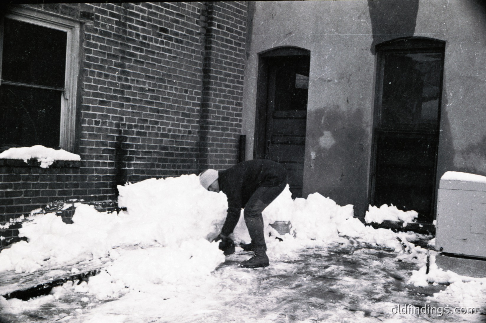 Mid-20th century black-and-white photo of a man clearing snow from a brick sidewalk. He wears a long coat, cap, and boots, shoveling snow into a pile beside a weathered brick building with arched windows and a door. Snow covers the ground and rooftops, indicating heavy winter conditions. The scene evokes post-WWII urban life.