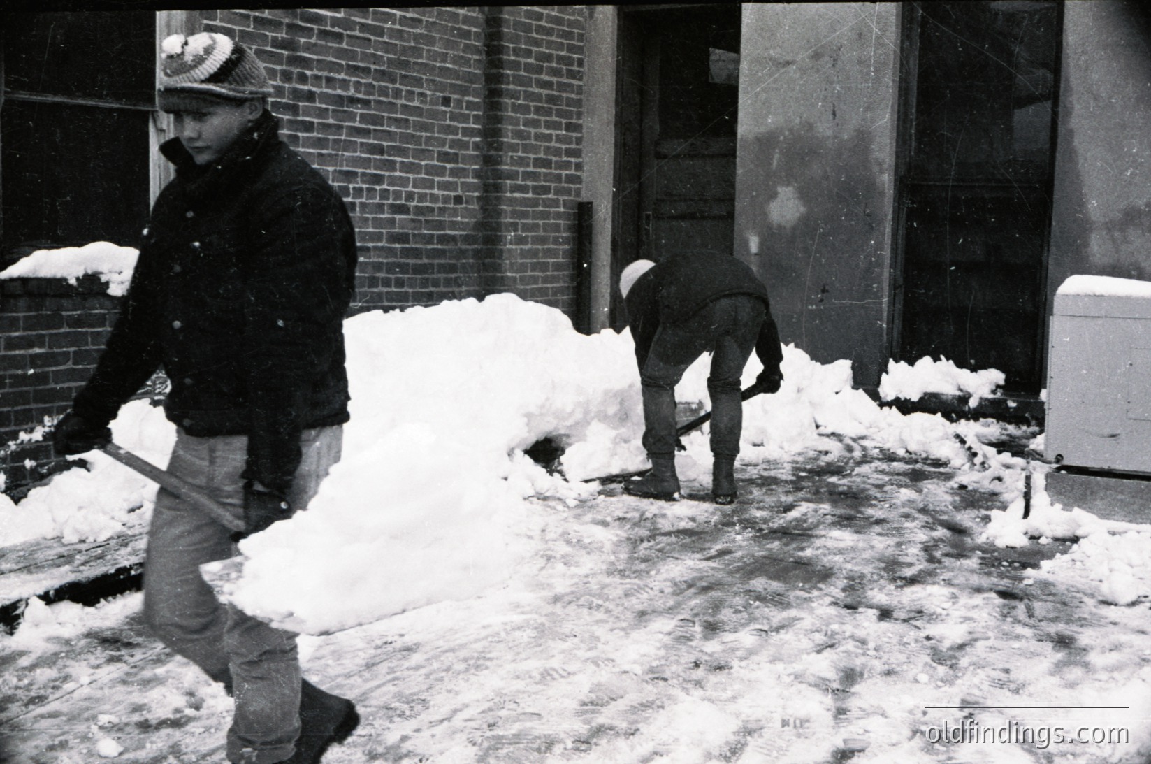 Two men clear snow from an urban sidewalk in mid-20th century, likely 1950s–1970s. One shovels while the other walks with a tool. Brick building and heavy snowfall suggest cold-weather city life.