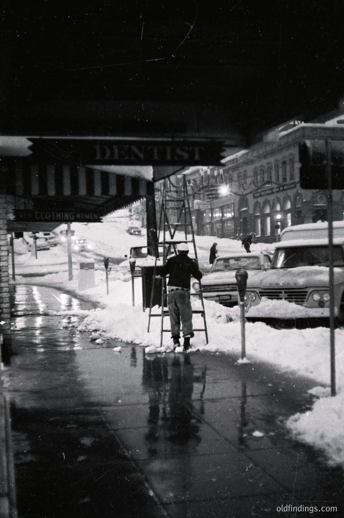 Mid-20th century urban street scene: worker clearing snow with ladder near a dentist’s office sign. Classic cars, snow-covered pavement, and vintage streetlights. Likely 1950s–1960s USA.