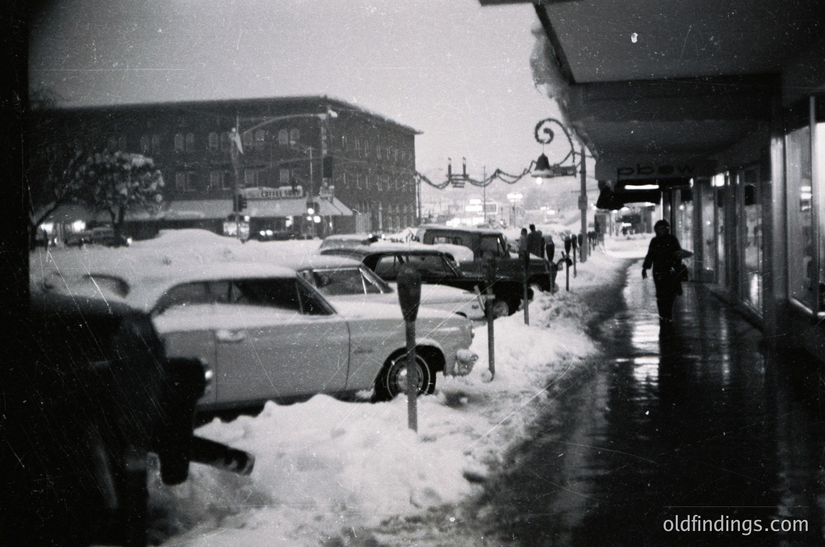 Vintage black-and-white street scene with mid-century cars parked along a snow-covered sidewalk. Classic 1950s–60s architecture features brick buildings and illuminated signs. Pedestrians bundled in winter coats navigate slushy streets. Snow-laden lampposts and hanging holiday lights add nostalgic charm.