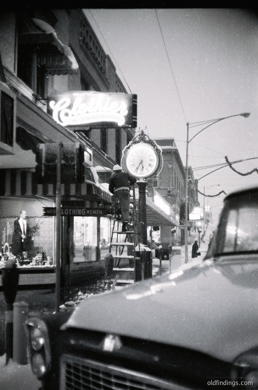 Mid-century street scene featuring a vintage clock pole with a round clock face displaying ~10:10. Classic storefronts with illuminated neon signs (#"Nothing" and a partial musical note symbol) line a quiet urban street. A parked vintage car (likely 1950s–1960s) frames the left edge. Architectural details include ornate storefronts and overhead wires.