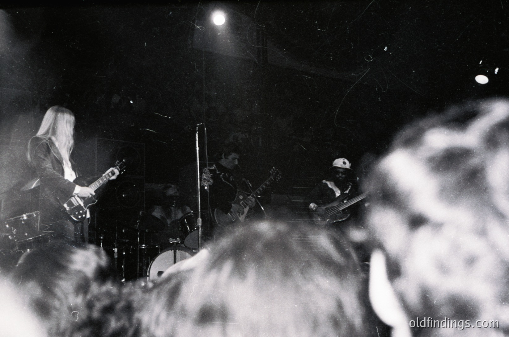 Three musicians performing live in dimly lit venue: guitarist (left), drummer (center), bassist (right). Crowd blurred in foreground. Black-and-white, high-contrast lighting. Likely 1970s–1980s rock/psychedelic scene.