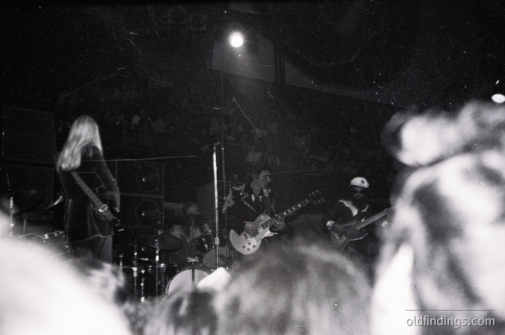 Intimate live band performance in dimly lit venue, featuring guitarist (left) and bassist (right) under spotlight. Crowd silhouetted in foreground, capturing raw energy of 1970s rock scene.