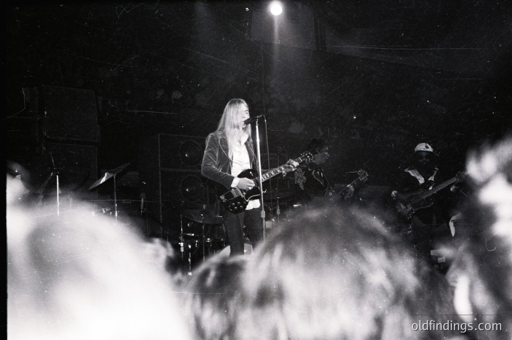 Guitarist performing live on stage, backlit by spotlight. Long hair and dark attire contrast against dark venue. Crowd silhouetted in foreground, capturing energy. Black-and-white, likely 1970s–1980s rock concert aesthetic. Authentic vintage concert vibe.