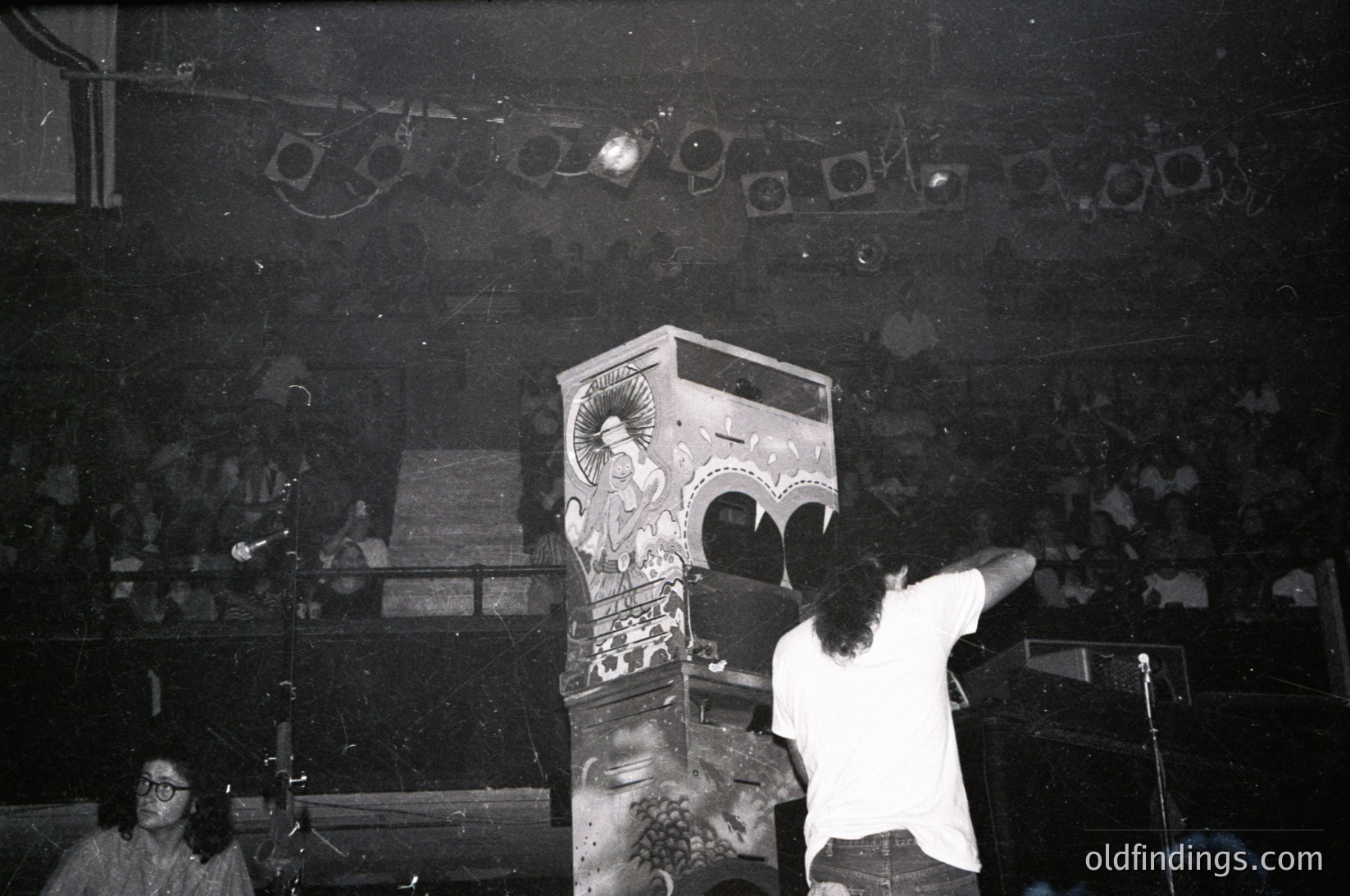 Vintage black-and-white concert shot featuring a DJ operating a large, retro arcade-style jukebox/music machine on stage. Crowd in dimly lit venue with stage lighting casting shadows. Artwork on machine depicts a stylized figure with tribal motifs. Likely 1970s–1980s underground music scene.