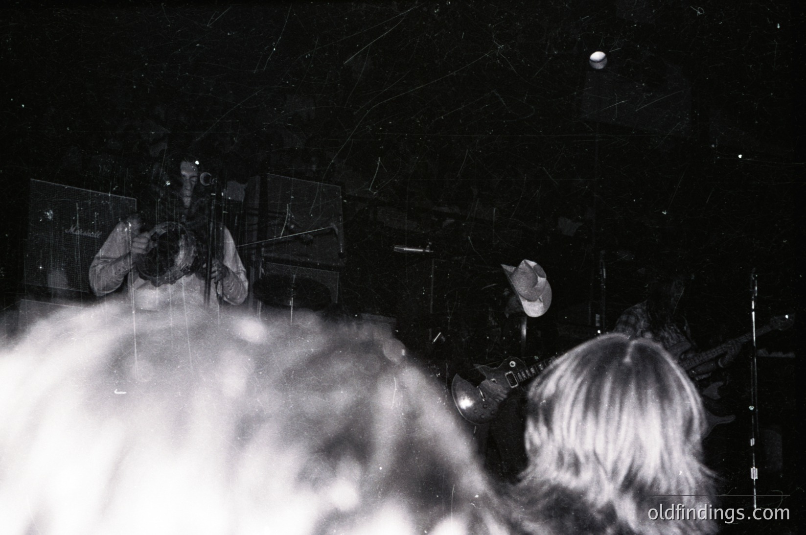 Vintage black-and-white shot of a live band performance in low-light, likely 1970s–1980s. Guitarist and drummer visible, with a spotlight illuminating the stage. Grainy texture and slight film damage enhance nostalgic atmosphere.
