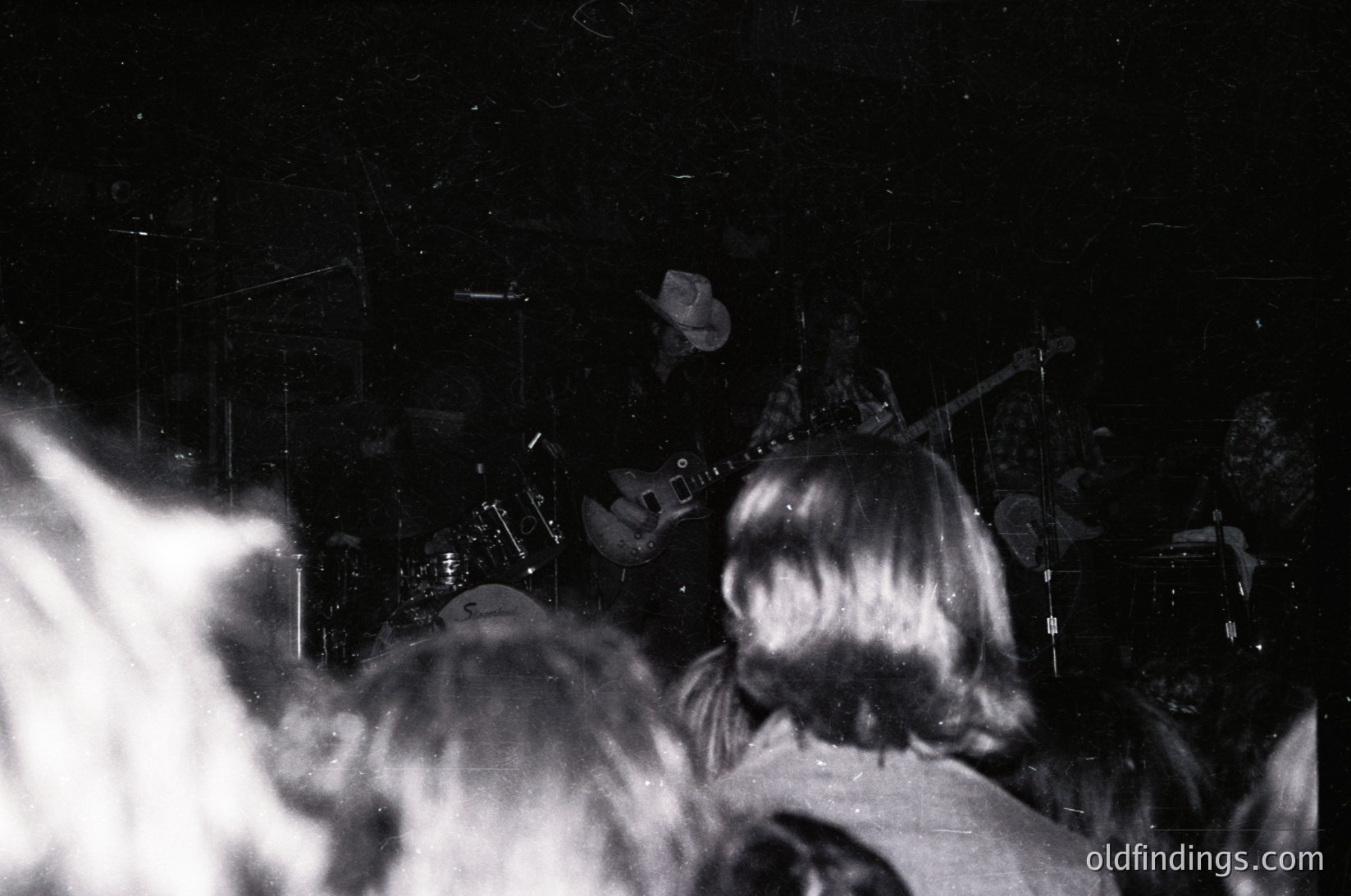 Black-and-white concert shot: stage lit by spotlight, guitarist playing electric guitar, drummer behind cymbals, audience silhouetted in dark venue. Likely 1970s–1980s rock/music scene.
