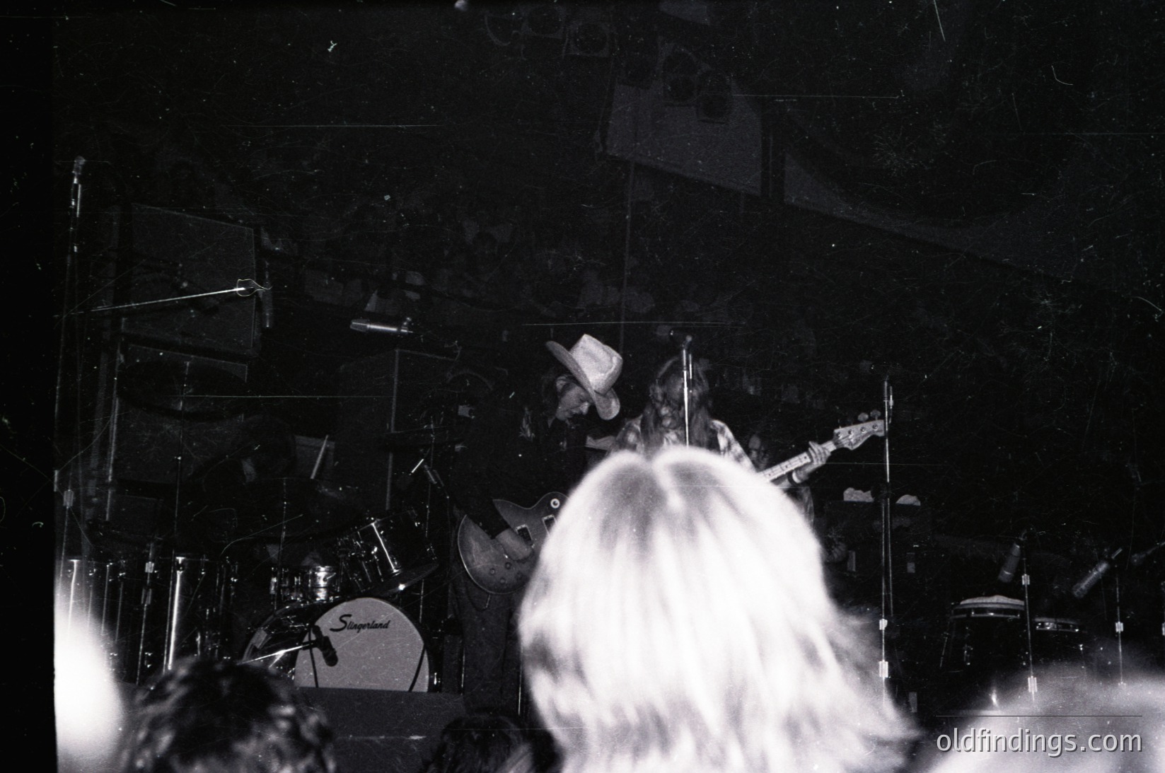 Black-and-white live performance shot featuring a drummer in mid-play with Slingerland drum set, surrounded by stage lighting and blurred audience. Captures raw energy of 1970s rock concerts.