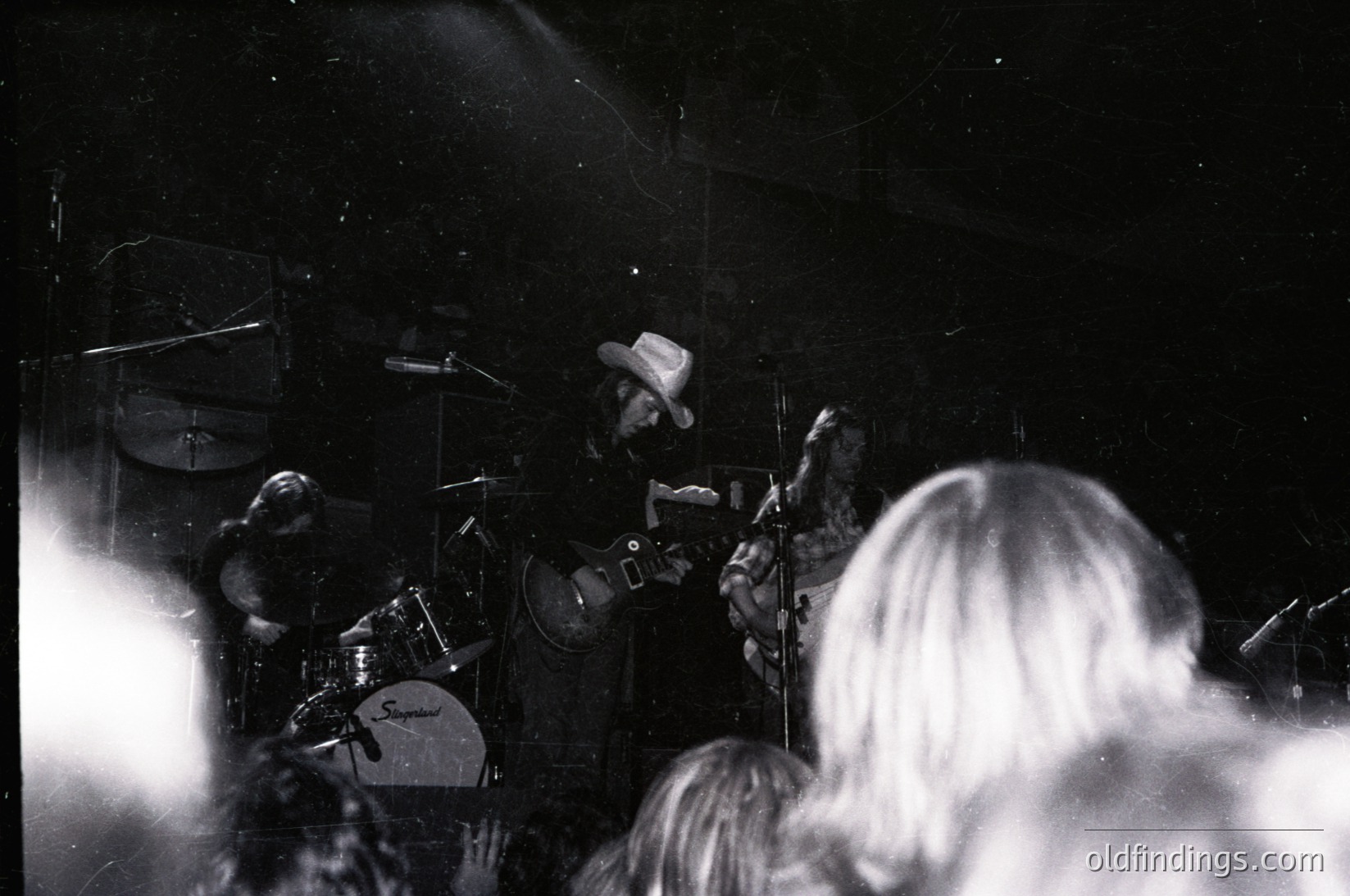 Intimate live blues/rock performance in monochrome, featuring guitarist (center) in fedora and leather jacket playing electric guitar, drummer (left) with cymbals and bass drum, and blurred audience facing stage. Stage lighting creates dynamic contrast. Style evokes 1960s–1970s rock clubs.