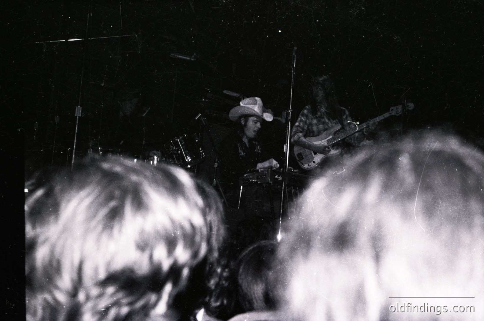 Blurred black-and-white concert shot: two musicians on stage—one playing drums (cowboy hat), one electric guitar—with audience silhouettes in foreground. Grainy, low-light aesthetic suggests live performance vibe. Likely 1960s–1970s folk/rock era.