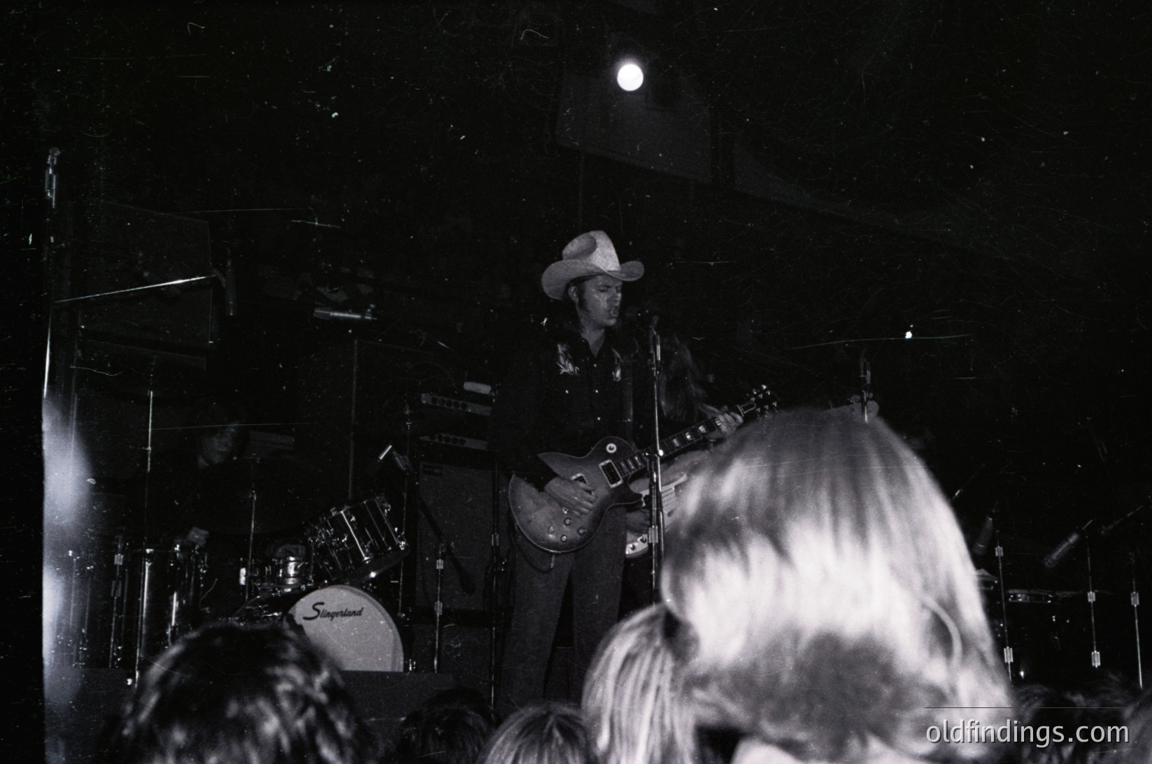 Black-and-white concert shot featuring a guitarist in cowboy hat playing a white electric guitar on stage. Stage lighting highlights the performer against a dark backdrop with drum set (Sperber brand) visible. Crowd silhouettes in foreground. Evokes 1960s–1970s live music era.